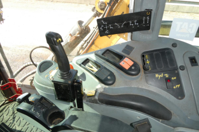 Tractor cab interior with joystick control, display panel, and various switches; visibility of hydraulic levers and a sticker with numbers on the window.