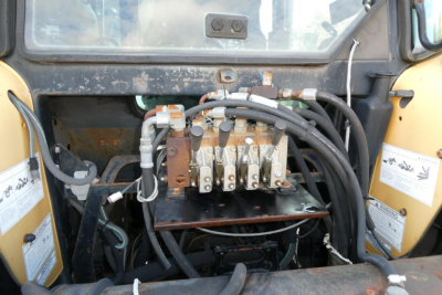 Tractor engine bay showing hydraulic control valves and hoses with metal manifold and orange-brown fittings, label stickers on sides, dusty interior.
