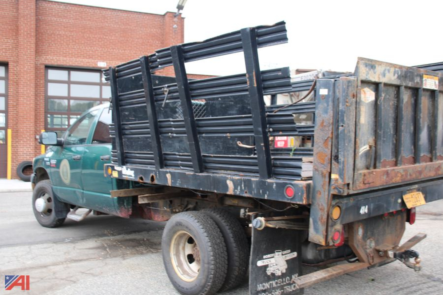 Dodge Ram 3500 dump truck with flatbed/rack and stacked metal rails; green cab, dark flatbed, rear view shows tailgate and equipment, image shows a work truck