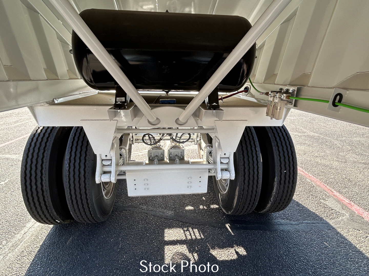 Rear view of a dump trailer showing dual rear axles and suspension components; undercarriage has a white painted frame and black tires, with mechanical linkage visible.