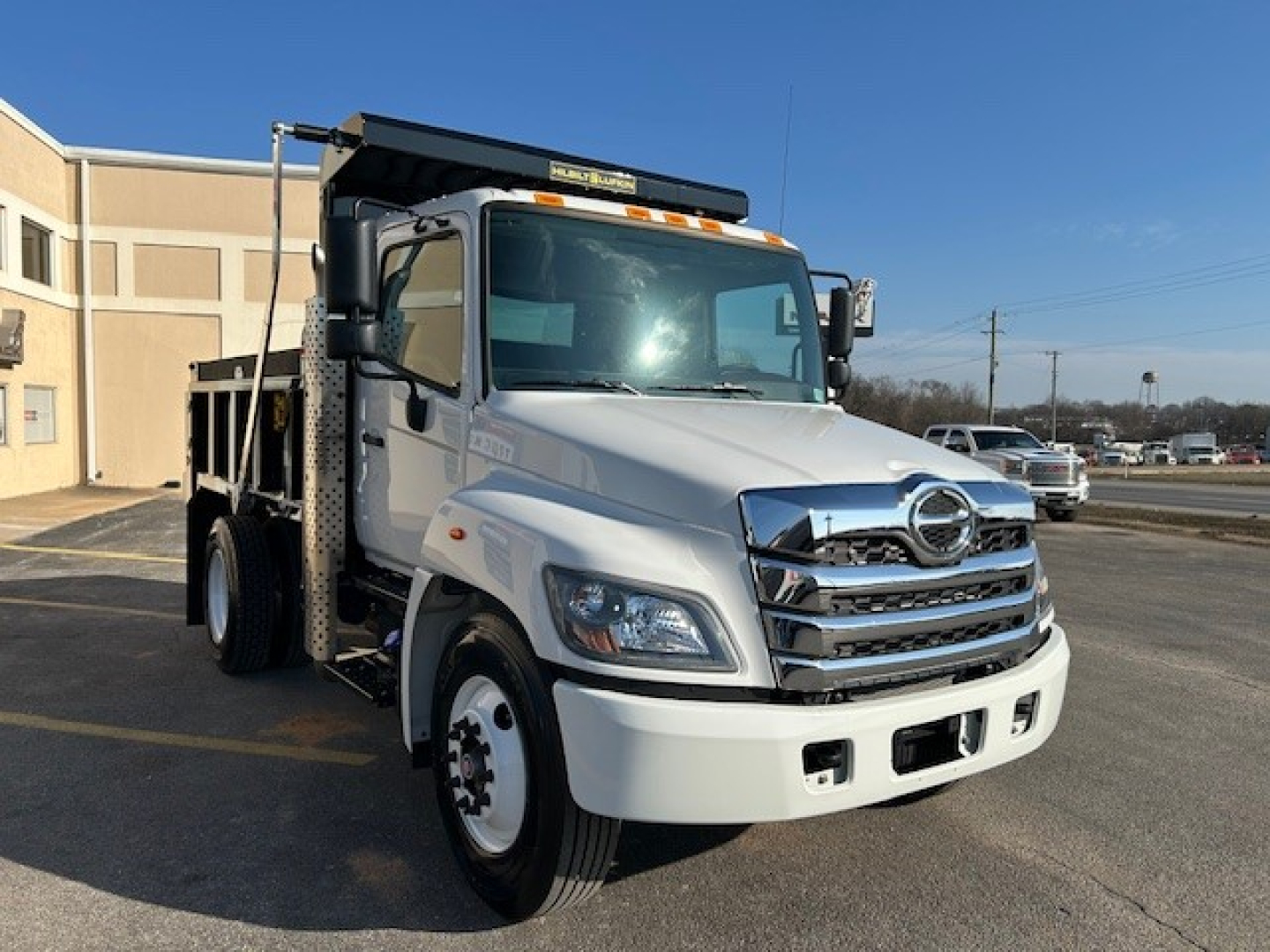 Hino L7 dump truck front-right view showing chrome grille, headlights, and dump body with black hoist; parked in lot.
