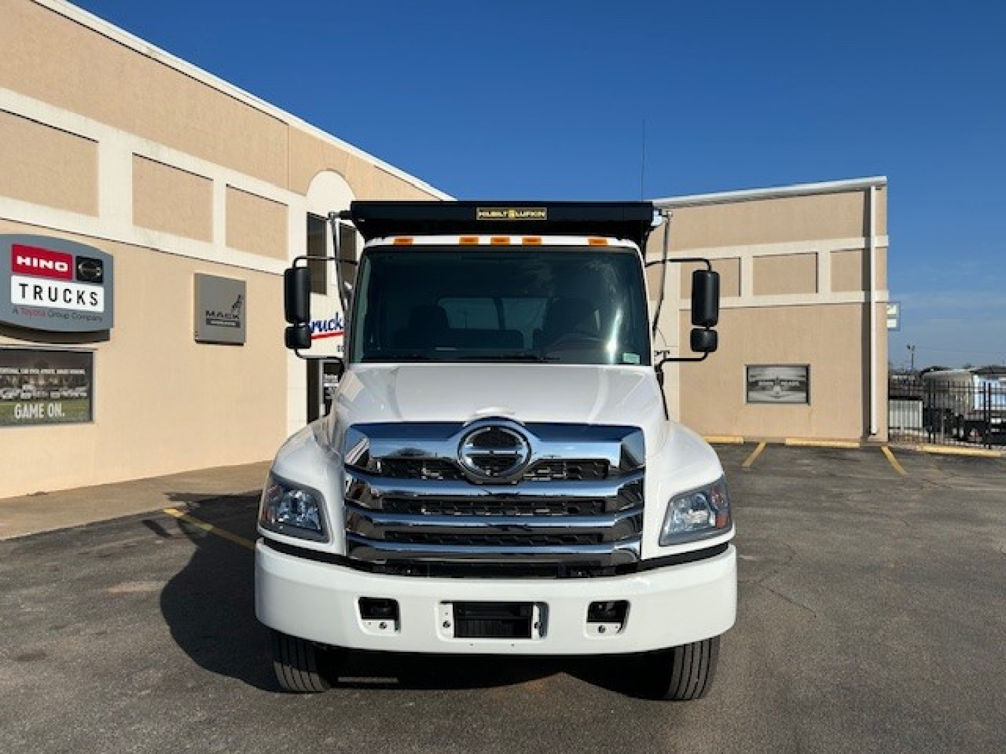 Front view of a white 2025 Hino L7 dump truck showing Hino Trucks logo on grille and dealership signage in background.