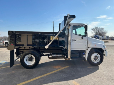 2025 Hino L7 dump truck side view showing dump bed with hydraulic mechanism, white cab, black dump body with Hino branding, large side mirrors, and wheel with white rim.