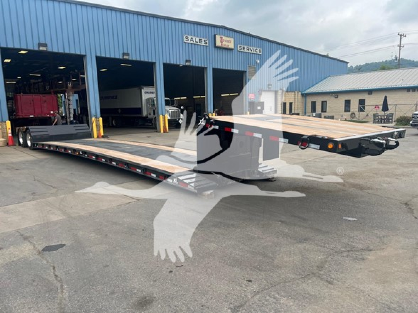 Lowboy trailer in front of blue service building showing long flatbed platform and ramps with wheels, id tag 53-6197, VIN 4U3J05324SL023562, appears used.