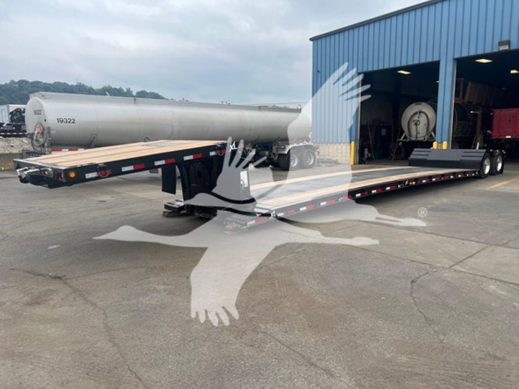 Lowboy trailer with long flat deck and gooseneck, attached to a towing vehicle. Tanker trailer in background, warehouse building to right.