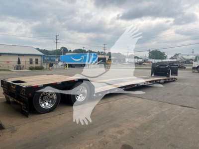 Lowboy trailer with long flatbed and multiple axles, visible wheels, hitch, and equipment in background.