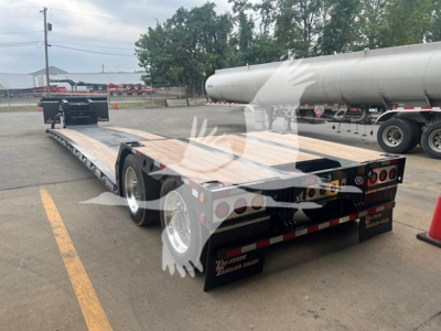 Trailer deck with lowboy flatbed showing wooden top, dual rear axles, and safety reflectors. Tanker truck in background and cones nearby.