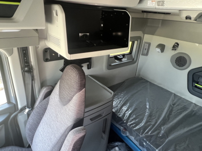 Interior cabin area of a 2026 Volvo VNL64T860 showing sleeper storage over cab, side shelf, and fabric seat with plastic-covered mattress in the background.