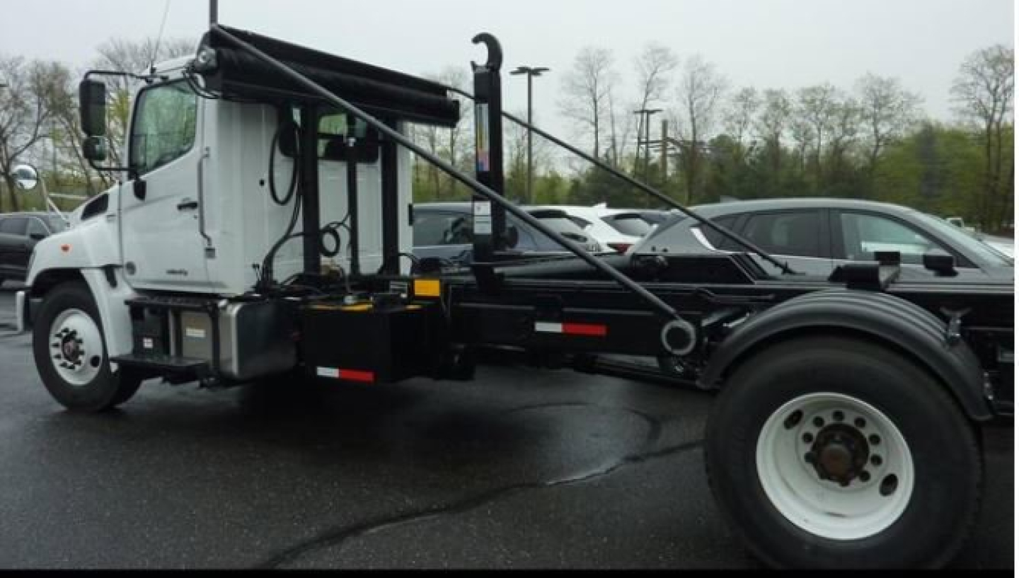 Hino L7 hooklift truck side view showing chassis frame, hydraulic hooklift mechanism, white cab with black hoist arms, front wheel, and parking lot background.