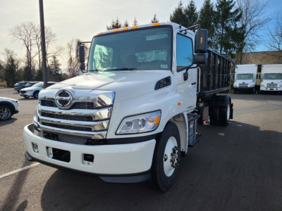 Truck cab front-left view of a white 2027 Hino L7 hooklift truck showing Hino emblem, chrome grille, headlights, side mirrors, and dump-bed in cargo area.