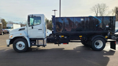 Truck side view showing a white cab and a large black dump bed on a hooklift truck chassis.