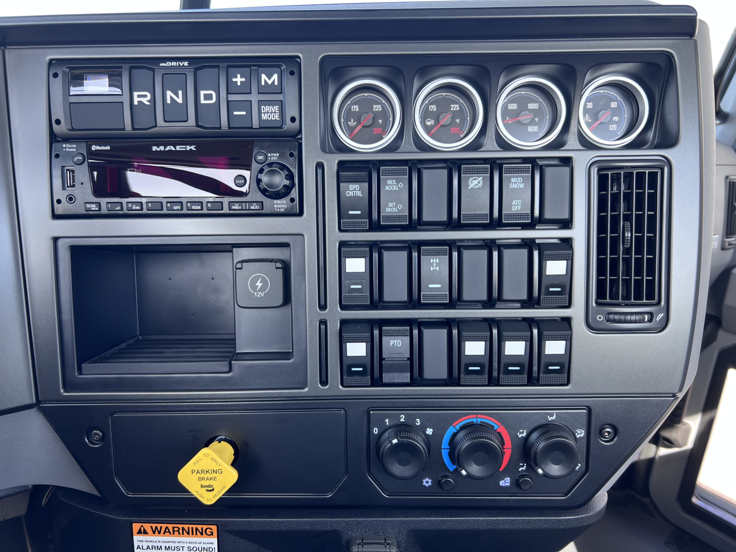 Dashboard view of a 2026 Mack GR64B cab and chassis showing gear selector, four round gauges, numerous switches, ignition, and climate controls.