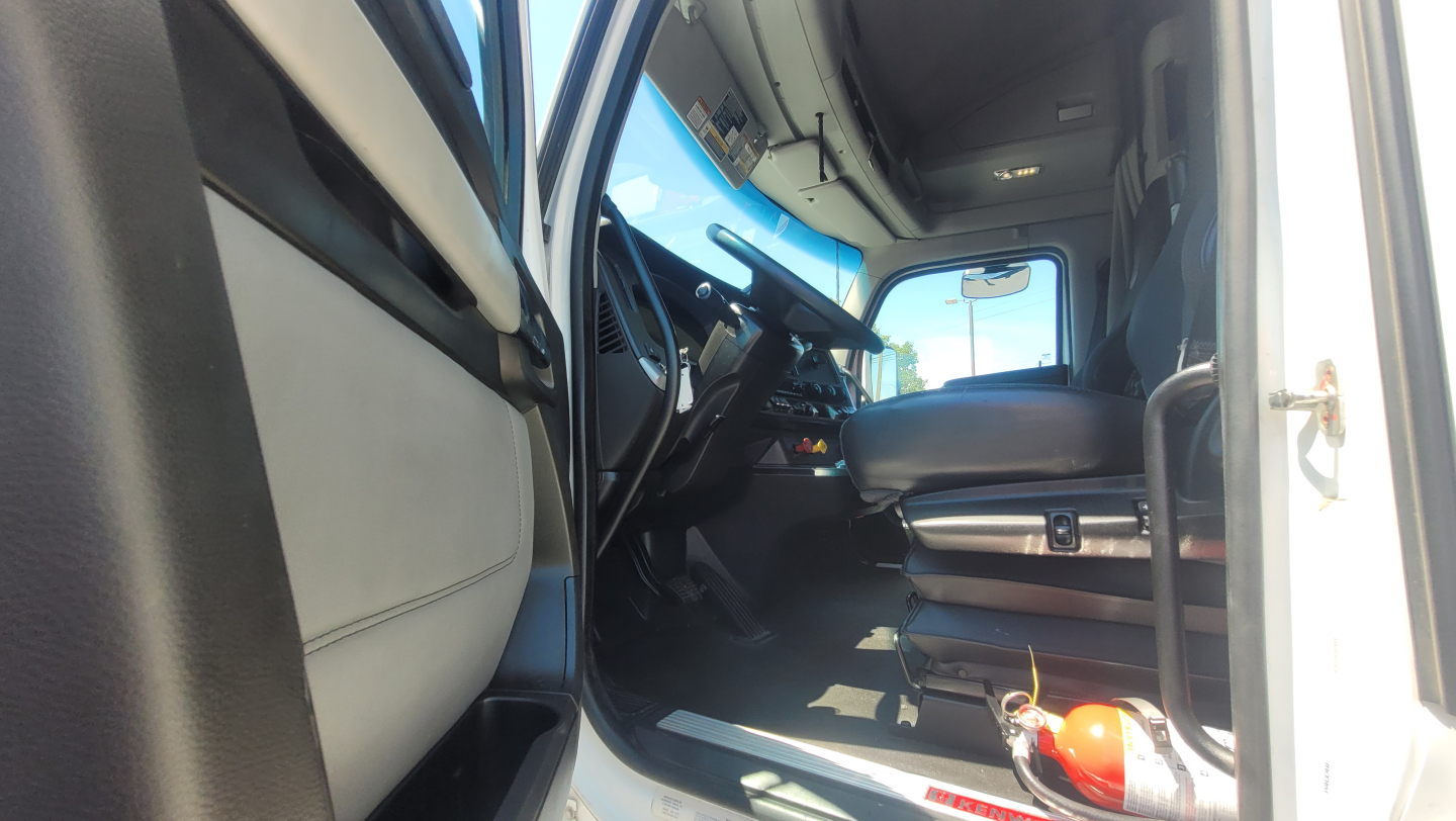 Interior of a Kenworth T680 truck cab showing driver's seat, steering wheel, dashboard, and entry door; visible are pedals and a fire extinguisher on the floor.