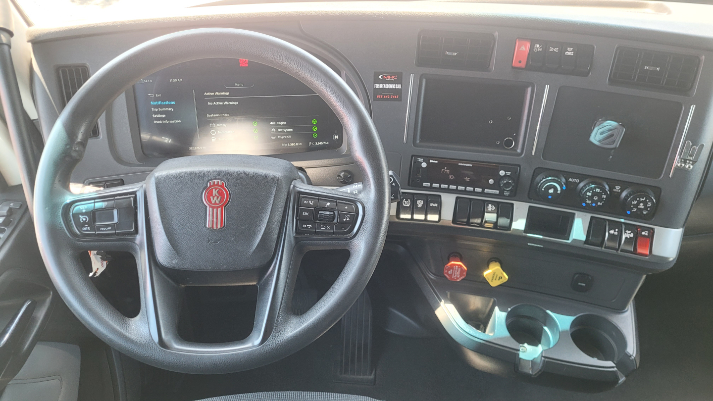 Kenworth T680 interior dashboard showing steering wheel with Kenworth emblem, digital gauges, center touchscreen, control switches, and cup holders.