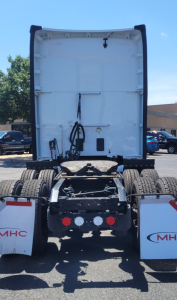 The back of a white Kenworth T680 tractor cab chassis showing the fifth wheel and rear mudflaps with 'MHC' branding. Seen are multiple rear tires, connectors, and wiring harnesses mounted on a trailer or tractor.
