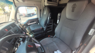 Interior of a Kenworth T680 cab showing driver and passenger seats, dashboard, and steering wheel; front seat fabric with Kenworth logo embossed on headrest.