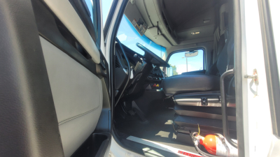 Interior of a Kenworth T680 truck cab showing driver's seat, steering wheel, dashboard, and entry door; visible are pedals and a fire extinguisher on the floor.