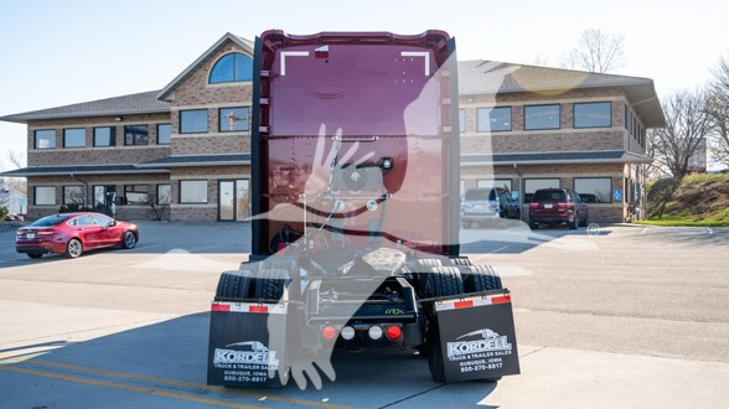 Kenworth T680 rear view showing wheel and sleeper cab, with trucks and building in background.