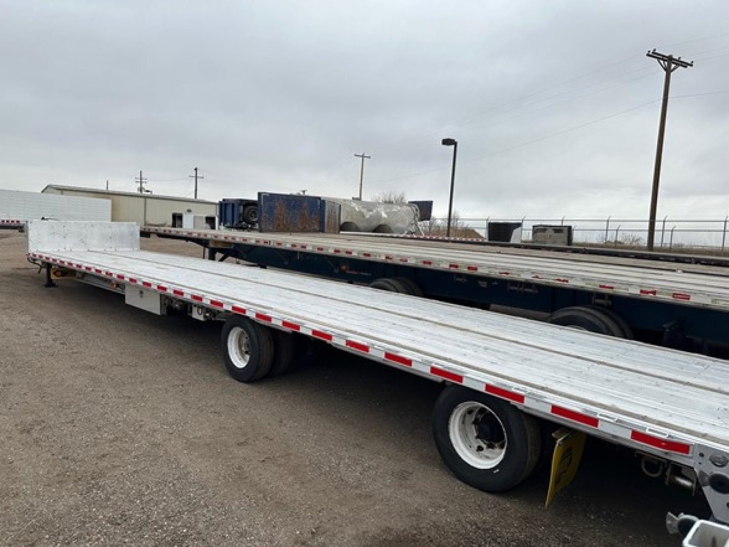 All-aluminum drop deck trailer showing long flatbed with side rails and dual rear axles, white aluminum deck, red/white reflective tape along edges.