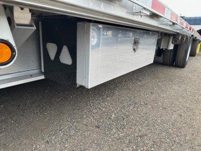 Drop deck trailer undercarriage showing aluminum toolbox mounted beneath deck and side frame with wheel and reflection in metal surface.