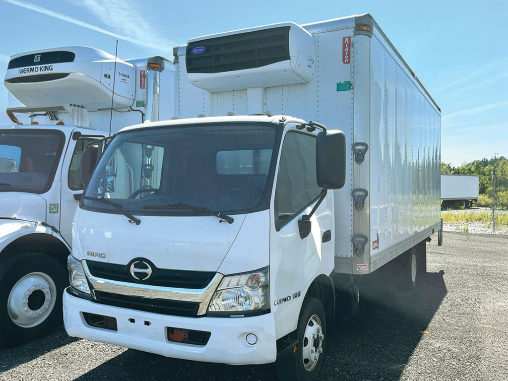Interior-freezer truck in-service white 2019 Hino 155 refrigerated box with cab in foreground, side mount refrigeration unit on top, other truck visible to left.