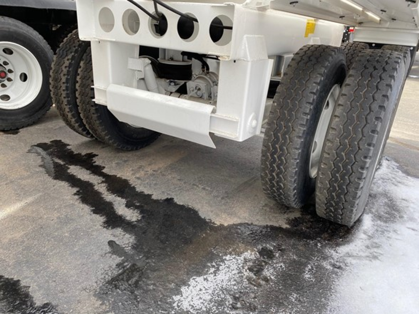 Trailer rear view showing dual rear axles and white chassis with heavy-duty suspension; tire treads visible; wet ground with patches of melted snow.