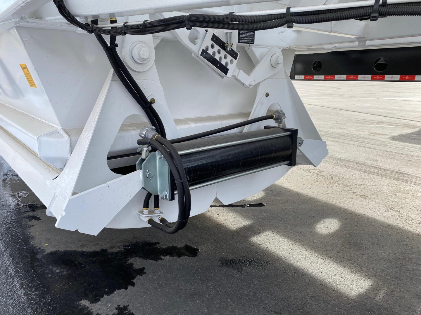 Dump trailer undercarriage showing winch/drum and hydraulic lines with white painted frame and black roller. Visible cables and mounting bracket.