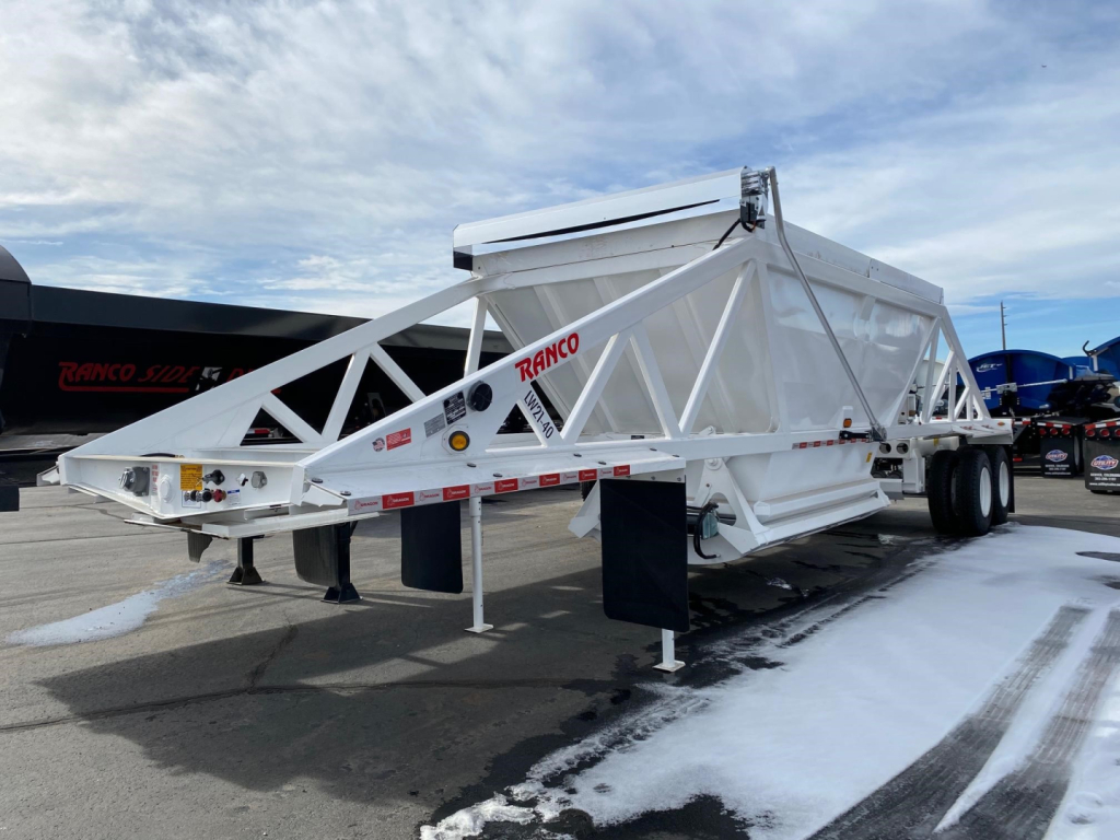Ranco LW21-40 dump trailer chassis in white with red RANCO branding, dual rear axles, and support jacks visible; side labels and hazard stripes along frame.