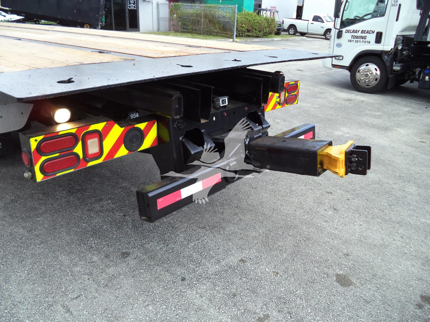 Truck rear showing towing hitch and bumper with red/yellow reflective decals, rear underride guard, black hitch receiver with yellow cap, and metal frame of a rollback bed.