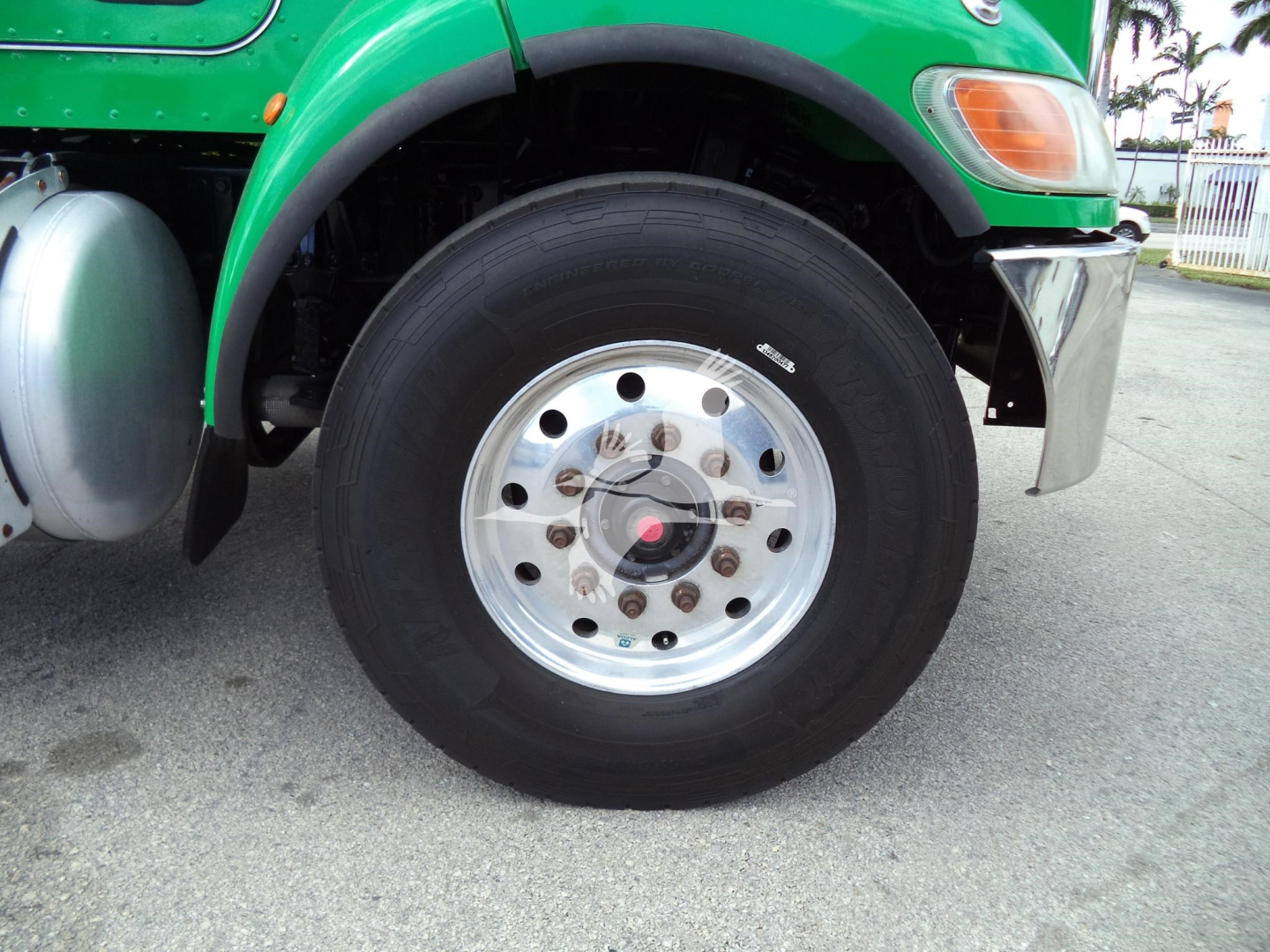 Green Peterbilt truck front wheel area showing silver alloy wheel with multiple lug nuts and black tire; visible text includes small white label on tire sidewall and chrome bumper nearby.