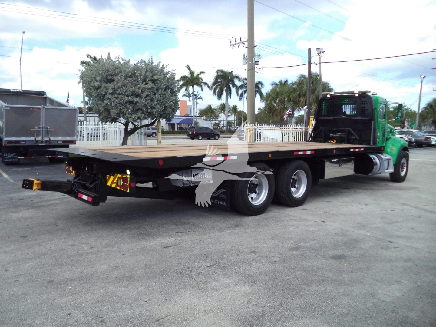 Peterbilt 348 rollback flatbed truck in a parking lot, long low-bed trailer attached, green cab, triple axles, rear view shows taillights and towing hitch.