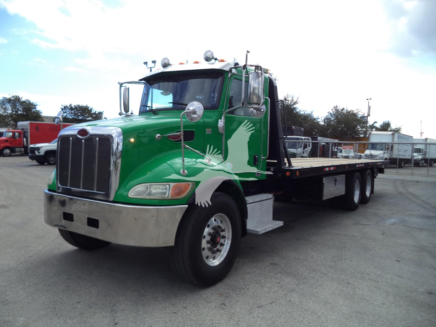 Peterbilt 348 rollback tow truck with a green cab and white angel wing graphics, flatbed body, chrome grille, dual rear axles, daytime roadside lot.