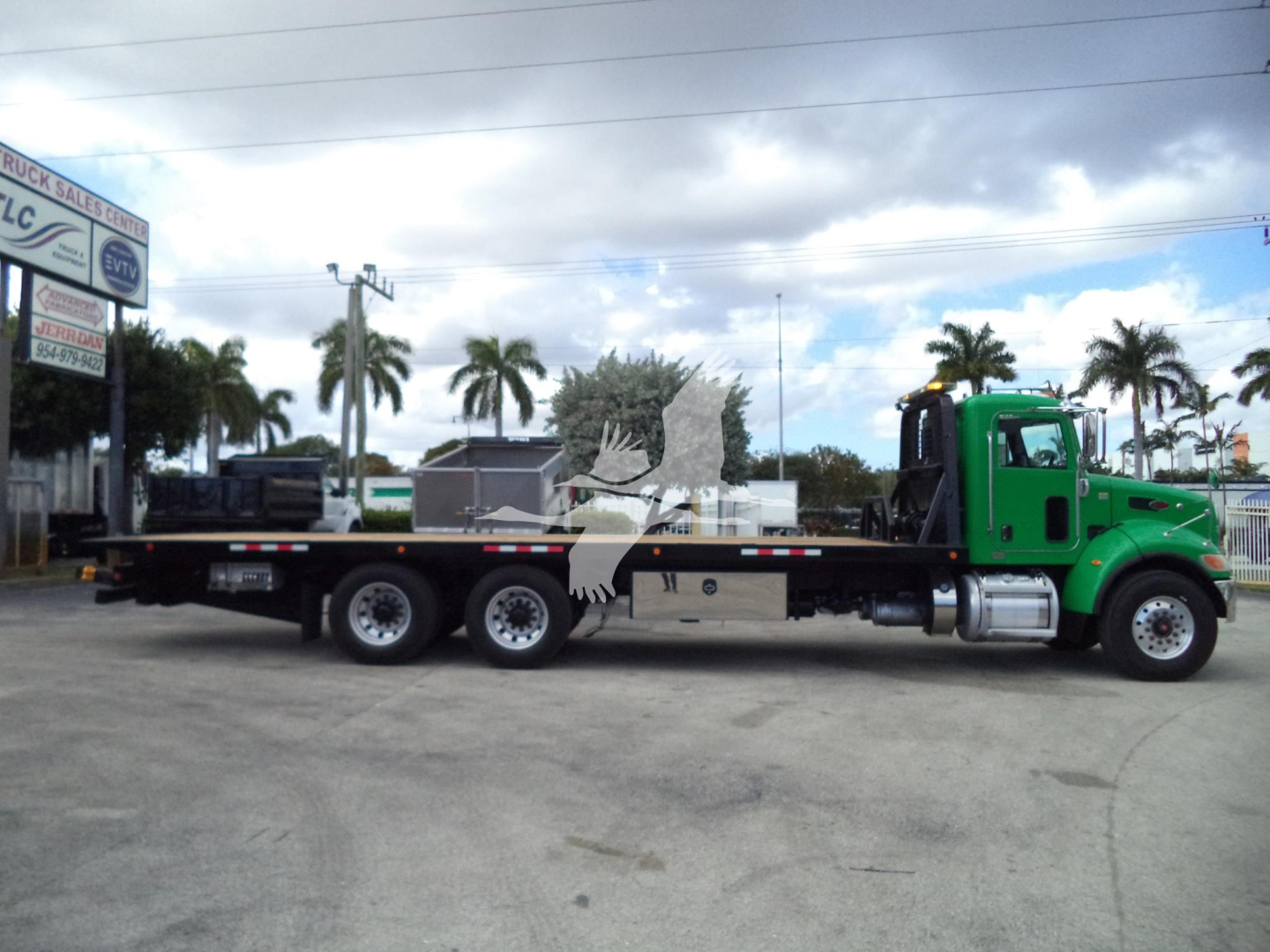 Peterbilt 348 rollback truck with green cab and long flatbed, three rear axles, dealership lot in background.