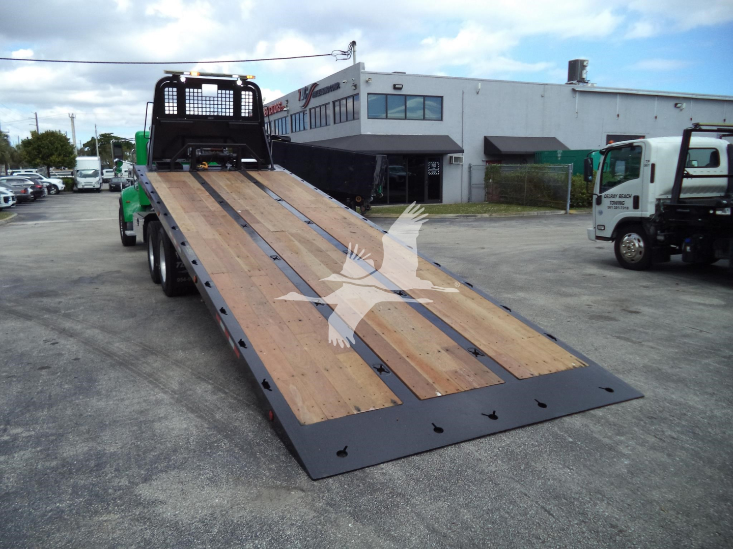 Rear view of a green Peterbilt rollback tow truck with a long wooden ramp, metal sides, and a black mesh cab guard. The bed is extended and there are other trucks and a building in the background.