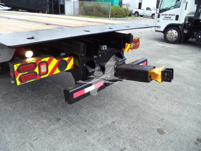 Truck rear showing towing hitch and bumper with red/yellow reflective decals, rear underride guard, black hitch receiver with yellow cap, and metal frame of a rollback bed.