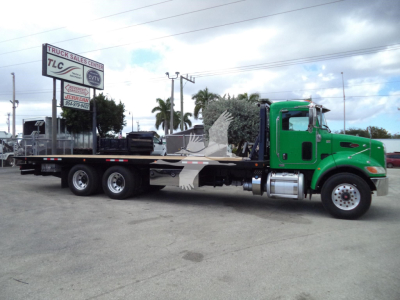Peterbilt 348 rollback truck with a green cab and long flatbed, parked at a lot; visible signboard and power lines in background.