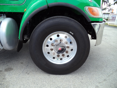 Green Peterbilt truck front wheel area showing silver alloy wheel with multiple lug nuts and black tire; visible text includes small white label on tire sidewall and chrome bumper nearby.
