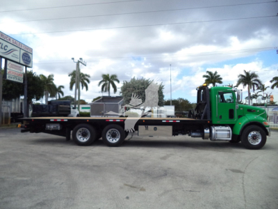 Peterbilt 348 rollback truck with green cab and long flatbed, three rear axles, dealership lot in background.