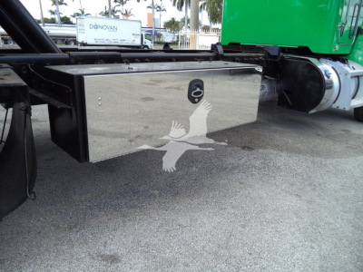 Trailer hitch/payload area with a shiny metal toolbox mounted under a Peterbilt chassis; view shows external tool storage box with rounded edges and a lock, on a rollback truck.