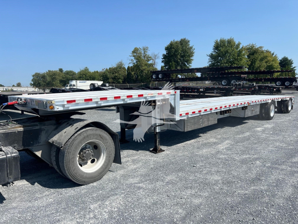 Flatbed trailer with drop deck and a heavy equipment transporter on a gravel lot. Long aluminum deck with reflective red/white safety stripes, dual rear axles, and wheel chocks.