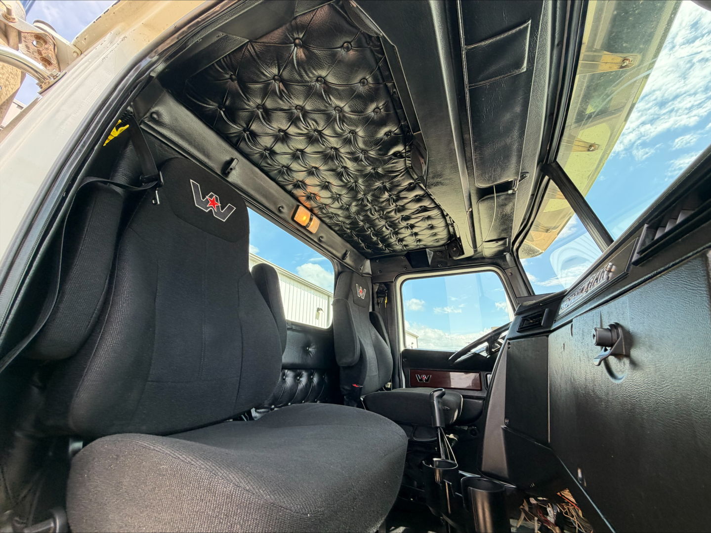 Cab interior of a 2019 Western Star 4900 SF with black quilted overhead liner, V-shaped seats with VTR logo, and basic dashboard; view from passenger side into cab showing seats and controls.