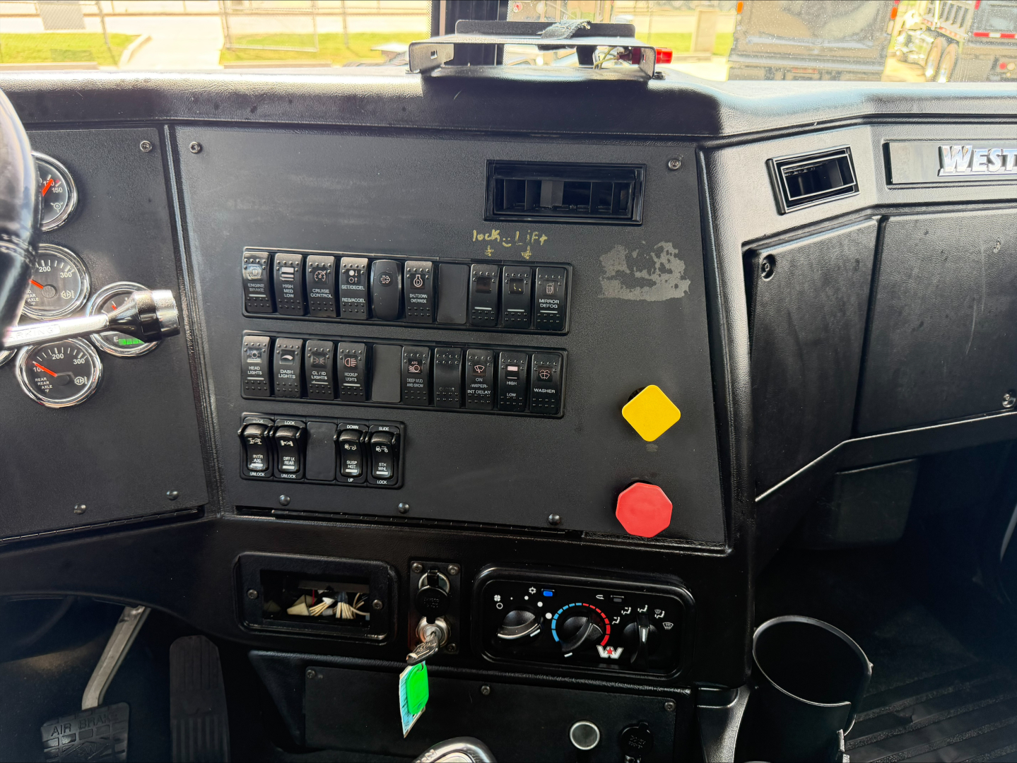 Truck cab dashboard with multiple switches and gauges, shows labeled switches and a green key tag below. The right side has a West logo and a black panel.