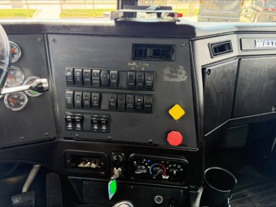 Truck cab dashboard with multiple switches and gauges, shows labeled switches and a green key tag below. The right side has a West logo and a black panel.