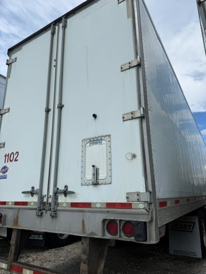 Rear view of a white utility trailer with double barn doors, steel hinges, and reflective red/white strips; small access door and bumper lights visible.