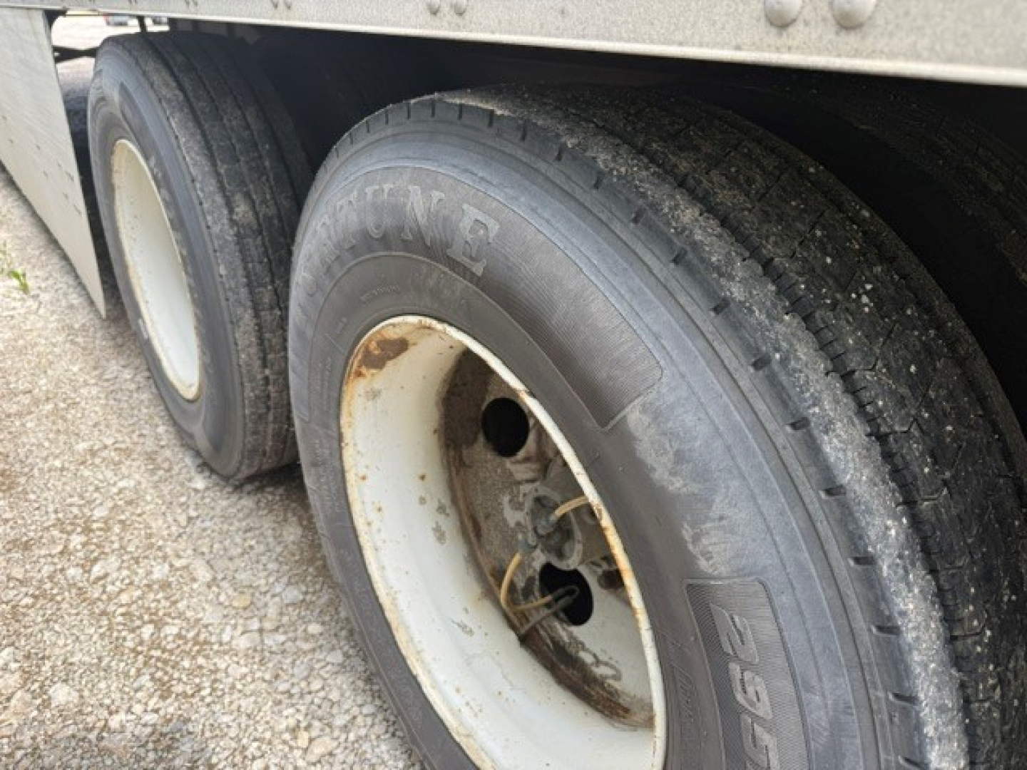 Trailer wheel showing Goodyear tire and white steel rim under utility trailer, with dual rear axle and mud/dust on surface.