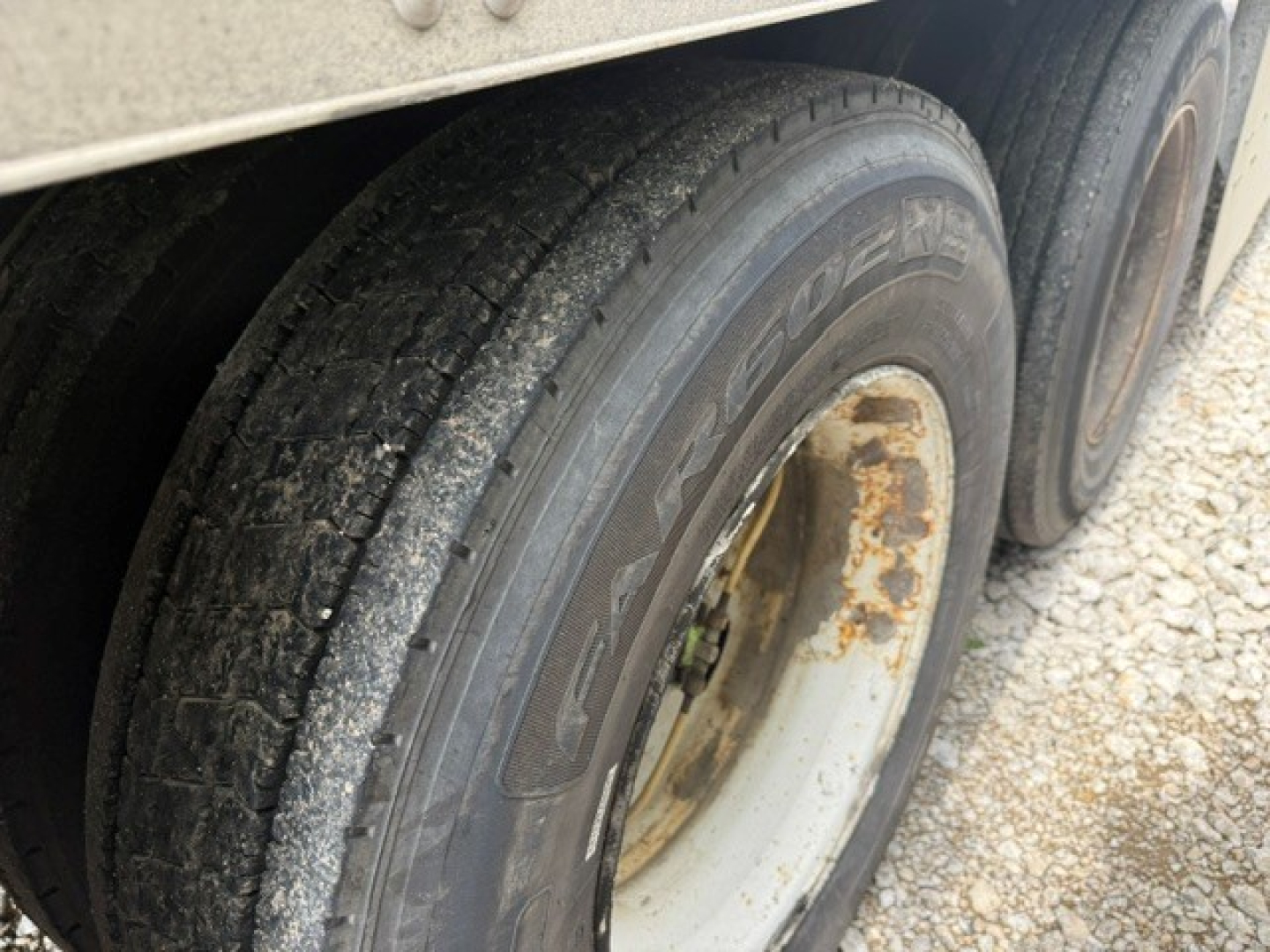 Trailer wheel close-up showing dual tires, tread and rim with rust on inner wheel; gravel ground.