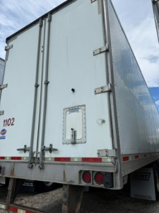 Rear view of a white utility trailer with double barn doors, steel hinges, and reflective red/white strips; small access door and bumper lights visible.