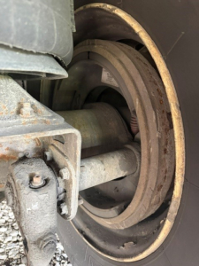 Trailer wheel hub and suspension components showing a large circular drum and metal arm with bolted bracket; rust-free looking metal surfaces; portion of tire and undercarriage visible.