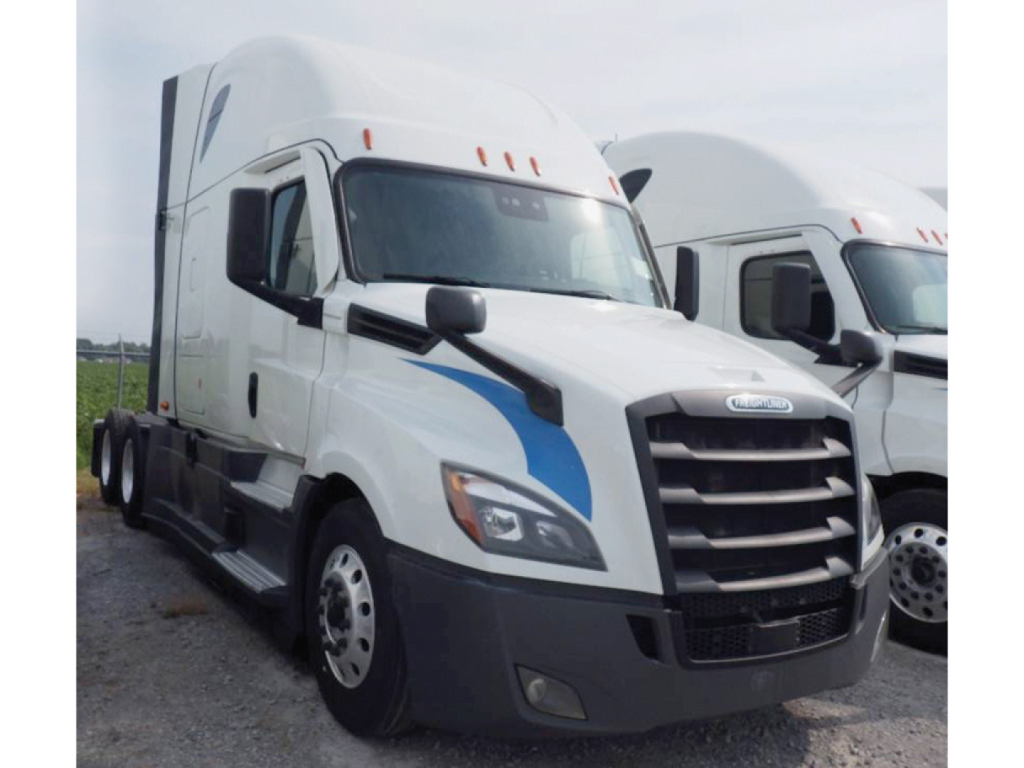 Truck cab showing 2020 Freightliner Cascadia126 with white exterior and blue graphic accents on the side, Daylight view, black grille, located in a gravel lot.