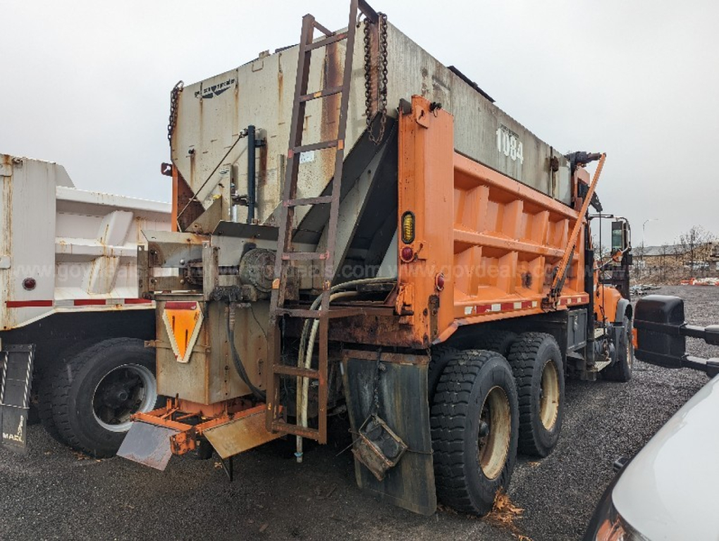 Mack CV713 Granite dump truck viewed from rear left with orange dump body and ladder, gravel bed, large tires, and rear hitch.